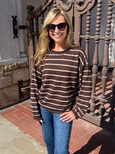 A woman wearing a brown and beige horizontally striped long-sleeve knit sweater, blue jeans, and dark sunglasses, standing outdoors in front of a decorative metal gate.