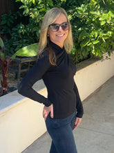 A woman with blonde hair and sunglasses wearing a black long-sleeve cowl neck top and dark blue jeans, standing outdoors on a concrete walkway with green foliage and a beige wall in the background.