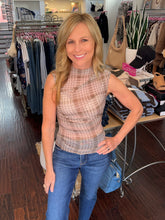 A woman with blonde hair stands in a clothing boutique wearing a sleeveless, high-neck plaid top in beige and brown tones with blue jeans.