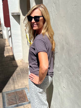 A woman with blonde hair wearing a dark gray t-shirt and light gray patterned pants, standing outdoors against a textured light gray wall.