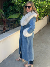 A woman wearing a long blue denim coat with a large white faux-fur collar and matching white faux-fur cuffs, standing outdoors on a concrete walkway with green plants in the background.