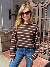 A woman wearing a brown and beige horizontally striped knit sweater, blue jeans, and dark sunglasses, standing outdoors in front of a decorative brown metal gate.