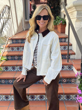 A woman wearing a white denim-style jacket with a dark brown collar, brown wide-leg pants, and a black-and-white striped shirt, sitting on outdoor tiled steps.