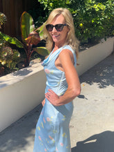 Woman in a light blue floral top, skirt and sunglasses standing outdoors with greenery in the background