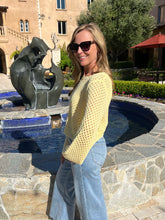 Woman in a yellow sweater and blue jeans standing in front of a pool with a sculpture in the background.