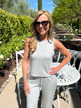 Woman in a gray outfit standing outdoors with greenery and a white chair in the background