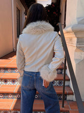 A woman viewed from behind wearing a light beige faux-leather jacket with a fluffy cream faux-fur collar and cuffs, paired with blue jeans, standing on outdoor terracotta steps.