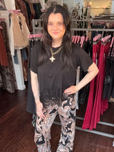 A woman with long dark hair stands in a clothing boutique wearing a black short-sleeve top with a sheer black lace hem and black, white, and gray marbled wide-leg pants. She is wearing a gold chain necklace with a cross pendant.