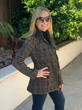 A woman wearing a brown and green plaid button-up shirt jacket over a black turtleneck, standing outdoors in front of a white wall and green foliage.