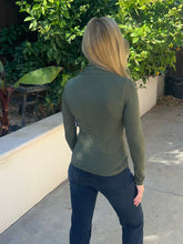 A woman viewed from behind wearing an olive green long-sleeve mock neck top and dark blue jeans, standing outdoors on a concrete patio with green plants and a white wall in the background.