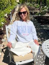 Woman sitting outdoors in a garden, wearing a 'Beach Bum' sweatshirt and sunglasses.