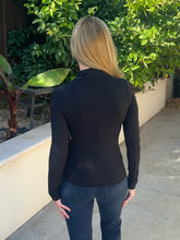 A woman viewed from behind wearing a fitted black long-sleeve top and dark blue jeans, standing outdoors on a concrete walkway with green plants and a beige wall in the background.
