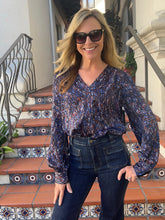 A woman wearing a long-sleeve V-neck blouse with a blue, purple, and orange abstract pattern, paired with dark blue jeans and sunglasses, standing on tiled outdoor stairs.