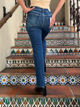 A person wearing blue denim jeans is shown from the back while standing on patterned tiled stairs. The photo focuses on the fit and back details of the jeans, with part of a beige top and black ankle boots also visible.