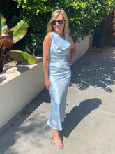 Woman in a light blue floral dress standing outdoors with greenery in the background