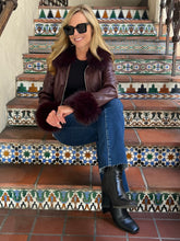 A woman sits on patterned outdoor stairs wearing a dark brown faux-fur trimmed leather jacket, blue jeans, black boots, and black sunglasses.