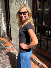 A woman with blonde hair wearing a dark charcoal gray v-neck t-shirt and blue jeans, standing outdoors in front of a building with a stucco wall and glass windows.