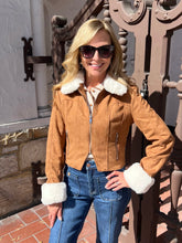 A woman wearing a cropped tan suede jacket with white faux-fur trim on the collar and cuffs, paired with blue jeans and sunglasses, standing outdoors in front of a decorative metal gate and stone wall.