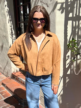 A woman wearing a tan suede cropped jacket, blue jeans, and sunglasses, standing outdoors against a stucco wall with shadows.
