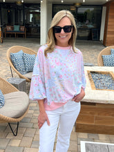 Woman in a pink patchwork top and white pants standing outdoors near patio furniture.