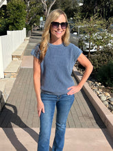 Woman wearing a blue ribbed knit top standing on a sidewalk with trees and cars in the background.