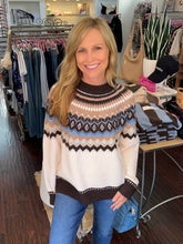 A smiling blonde woman wearing a cream, brown, beige, and light blue Fair Isle patterned knit sweater with blue jeans, standing inside a clothing boutique.