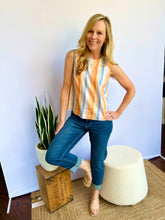 Woman wearing a striped sleeveless top and jeans standing in a room with a plant and ottoman.