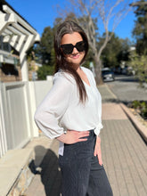 Woman wearing sunglasses and a white blouse with black pinstripe pants outdoors.