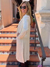 A woman wearing a long cream-colored blazer and sunglasses stands on outdoor terracotta tiled steps in front of a building with a stone column and black metal railing.