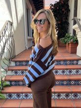 A woman wearing a brown and light blue striped off-the-shoulder sweater and brown pants, standing on tiled outdoor steps in front of a building.