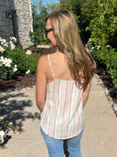 Woman wearing a striped tank top and sunglasses, standing on a sidewalk with greenery and a building in the background.