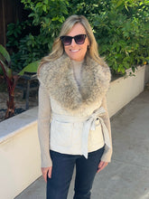 A woman wearing a cream quilted wrap vest with a large beige faux fur collar, styled with a beige knit sweater, dark pants, and sunglasses, standing outdoors on a concrete walkway with green plants in the background.