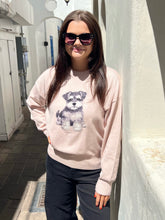 A woman wearing a light pink crewneck sweatshirt with a dog graphic on the front, paired with dark pants and sunglasses, standing outdoors against a white wall.