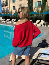 Woman in a red sweater and denim shorts standing by a poolside.