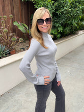 A woman wearing a light gray ribbed mock neck sweater and dark gray pants, standing outdoors on a concrete walkway with green plants and a wooden fence in the background.