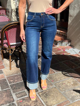 A person wearing dark blue straight-leg jeans with rolled cuffs, paired with a beige top and clear high-heel sandals, standing on a stone patio.