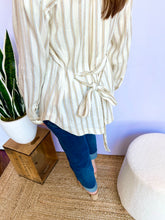 Woman wearing a striped shirt and blue jeans standing next to a potted plant against a light blue wall.