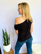 Woman wearing a black off-shoulder top and blue jeans standing against a white wall with a plant on a small table to her left.