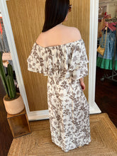 Woman in a floral off-shoulder top with matching pant holding showing the back view, standing against a wooden panel wall.