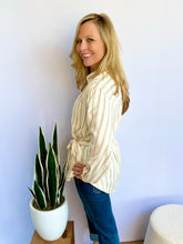 Woman wearing a striped shirt and blue jeans standing next to a potted plant against a light blue wall.