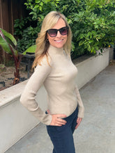 A woman wearing a beige ribbed mock neck sweater and dark sunglasses, standing outdoors in front of a planter with green foliage.