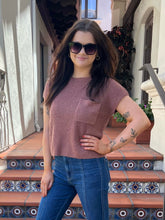 A woman wearing a dusty rose ribbed short-sleeve top with a chest pocket and blue jeans, standing on tiled outdoor steps.