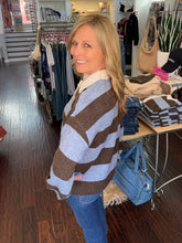 A woman with blonde hair wearing a brown and light blue striped knit sweater and blue jeans, standing inside a clothing boutique.