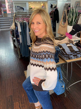 A woman posing in a clothing boutique wearing a knit sweater with a brown, beige, and light blue Fair Isle pattern over a white base, paired with blue jeans.