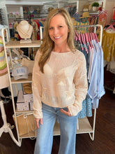 Woman wearing a beige sweater and blue jeans standing in a store with clothing racks and shelves in the background.