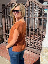 A woman wearing a short-sleeve ribbed knit top in a burnt orange color, paired with blue jeans and sunglasses, standing outdoors in front of a decorative brown metal gate.