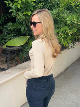 A woman with blonde hair and sunglasses, wearing a beige ribbed long-sleeve top and dark blue jeans, standing outdoors in front of a green hedge and a beige wall.