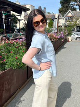 Woman in sunglasses and light blue shirt standing outdoors with flowers and buildings in the background