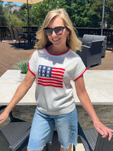 Woman wearing a white sweater with an American flag design outdoors.