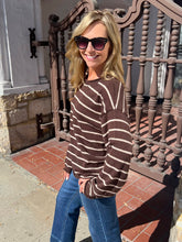 A woman wearing a brown and cream striped knit sweater, blue jeans, and sunglasses stands on an outdoor porch with a decorative brown railing.